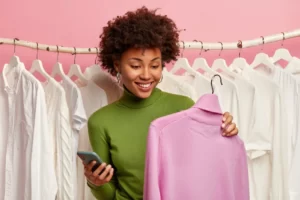 side-hustle positive-black-woman-chooses-sweater-buy-holds-hanger-with-purple-turtleneck-mobile-phone-other-hand