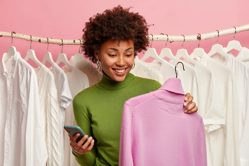 positive-black-woman-chooses-sweater-buy-holds-hanger-with-purple-turtleneck-mobile-phone-other-hand